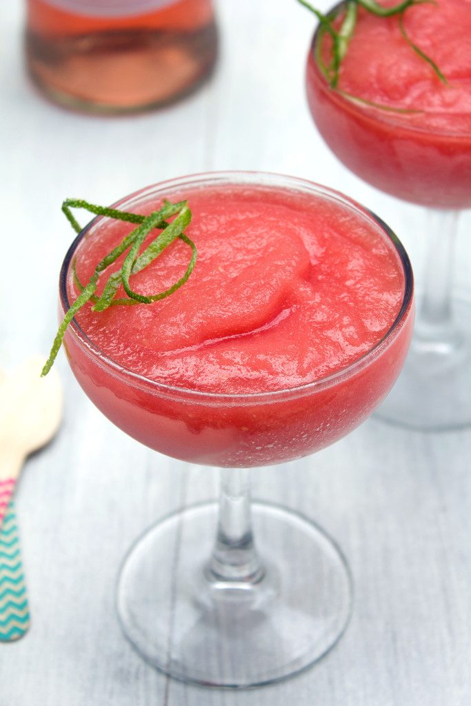 Head-on closeup view of watermelon rosé slushie with second glass in the background, wooden spoons, and bottle of rosé