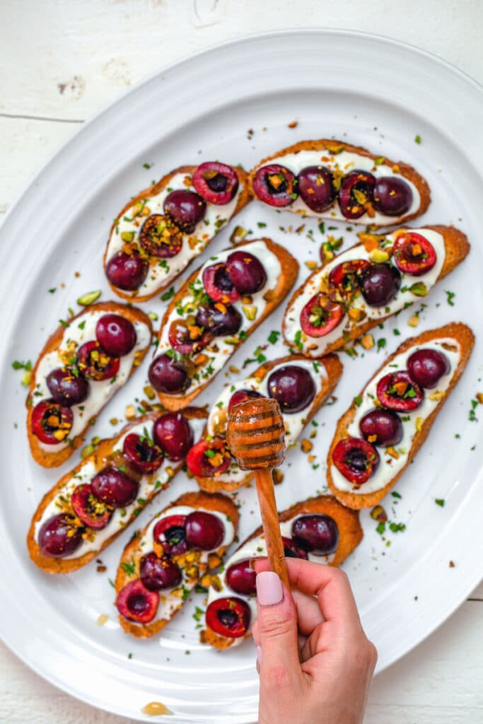 Overhead view of white platter with whipped ricotta cherry crostini with hand holding honey comb drizzling honey over them