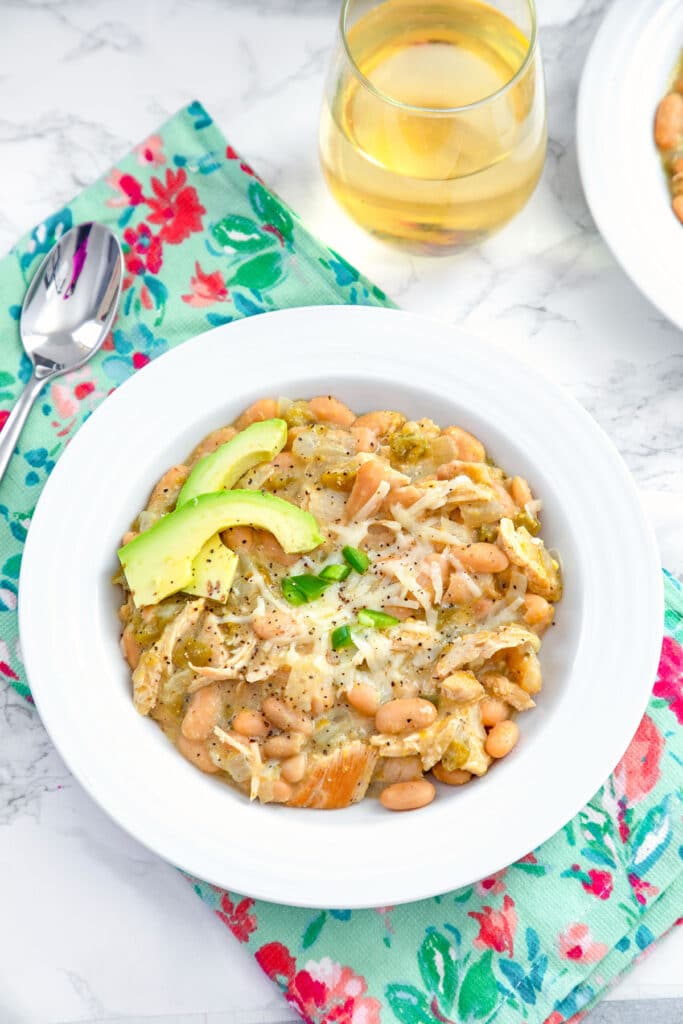 Overhead view of a bowl of white chicken chili topped with cheese, avocado, and jalapeño with spoon and glass of white wine in the background.