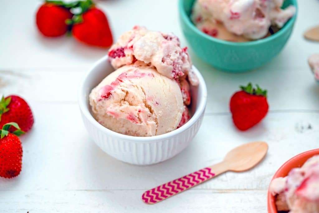 Landscape view of white bowl of white chocolate strawberry ice cream with other filled bowls in background, whole strawberries, and pink chevron wooden spoon