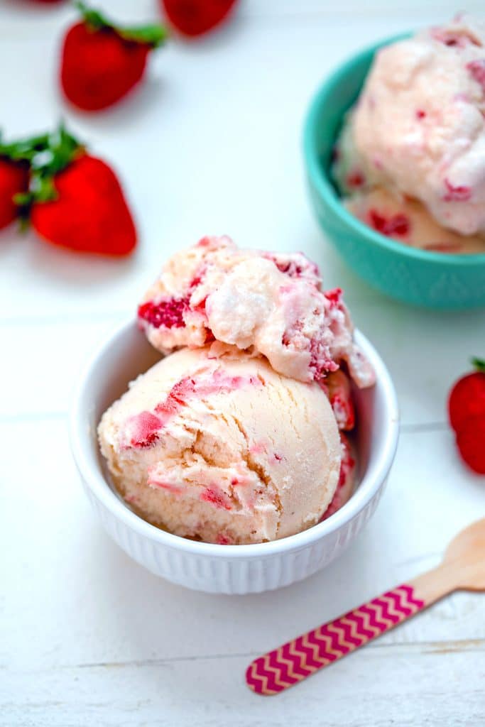 Overhead view of white chocolate strawberry ice cream in a white bowl with lots of strawberries in the background, pink chevron wooden spoon, and second teal bowl of ice cream