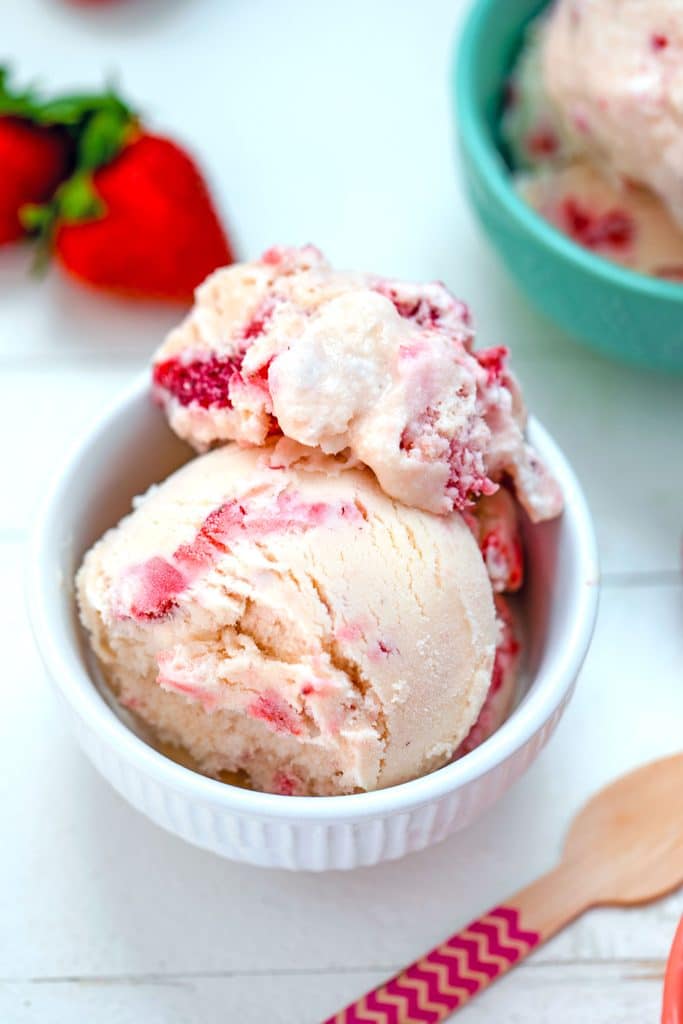 Overhead view of white bowl of white chocolate strawberry ice cream with strawberries, pink wooden spoon, and second bowl in the background