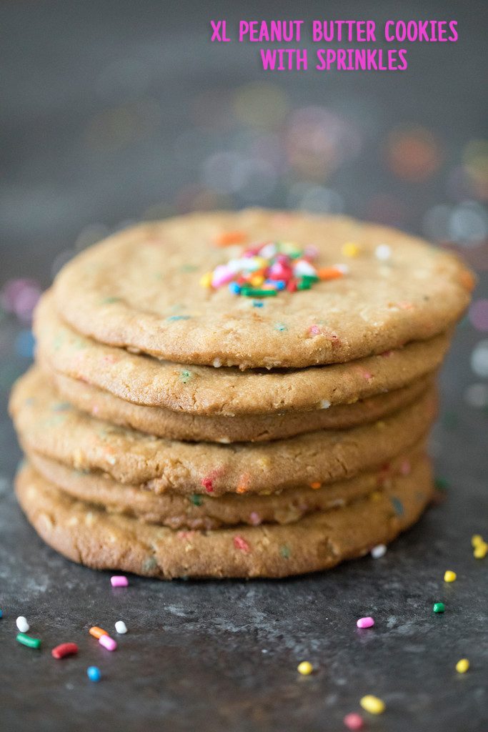 Head-on view of a stack of XL peanut butter cookies with sprinkles on a dark surface surrounded by sprinkles with recipe title at top