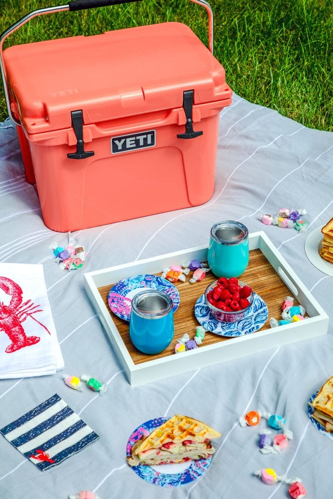 Close-up of picnic spread featuring coral Yeti cooler on a grey blanket with a tray with blue cups, lobster napkins, and plates with waffles and waffles sandwiches