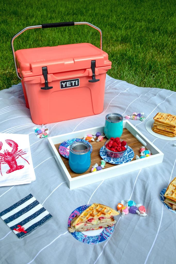 Picnic spread featuring coral Yeti cooler on a grey blanket with a tray with blue cups, lobster napkins, and plates with waffles and waffles sandwiches