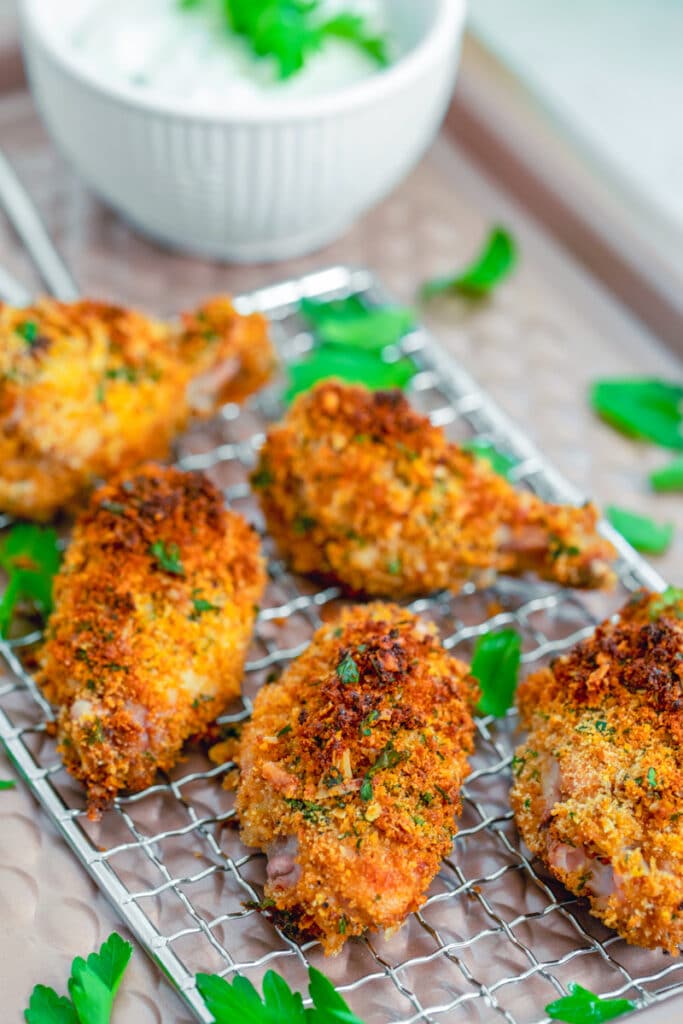View from a distance of a baking rack with golden zesty baked chicken wings surrounded by chopped parsley with bowl of yogurt dipping sauce in the background