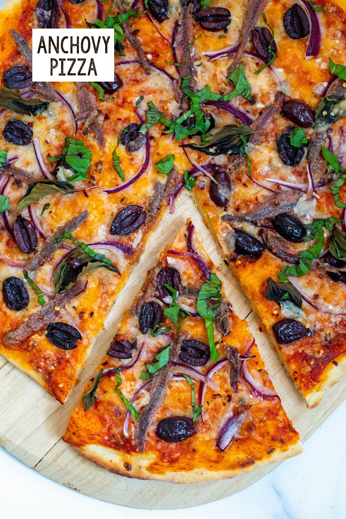 Overhead view of a pizza with anchovies, tomato sauce, red onion, black olives, and basil with a slice pulled out from rest of pizza and recipe title at top.