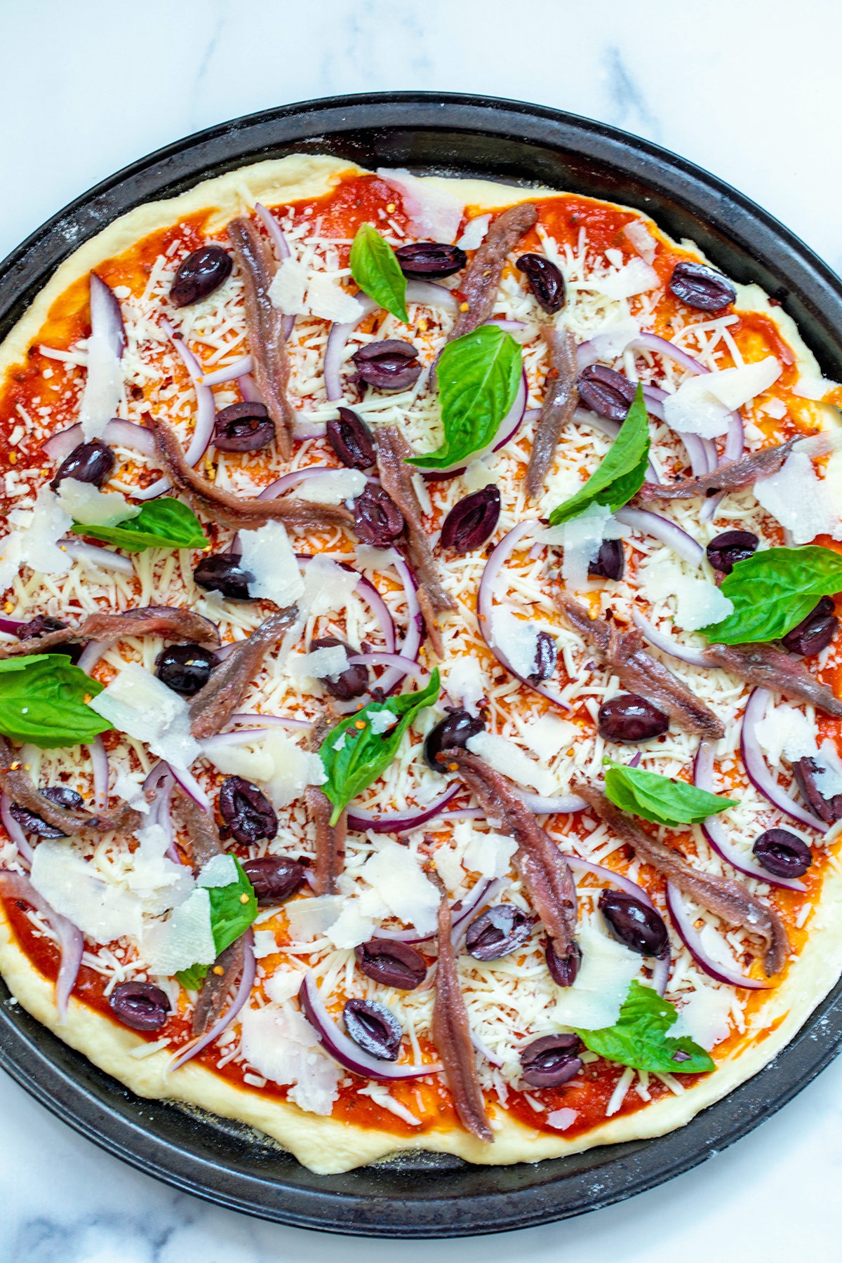 Pizza dough spread out on pan with tomato sauce, shredded cheese, shaved parmesan, anchovies, olives, red onion, and basil leaves.