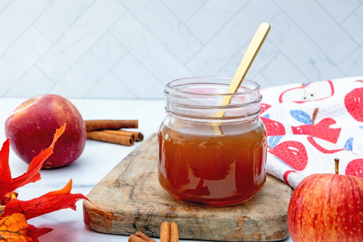 Landscape head-on view of a small jar of apple cider syrup with spoon with apples and cinnamon sticks in background.