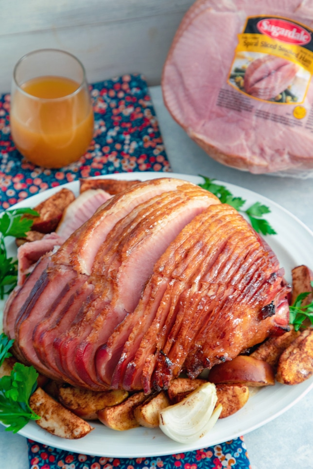 Vue de dessus d'un jambon glacé aux pommes et aux épices coupé en spirale sur un plateau entouré de pommes au four et de quartiers d'oignons avec un verre de cidre de pomme et un autre jambon en arrière-plan