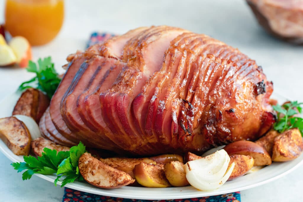 Head-on landscape view of spiral sliced apple spice glazed ham on a white platter surrounded by baked apples, onions wedges, and parsley