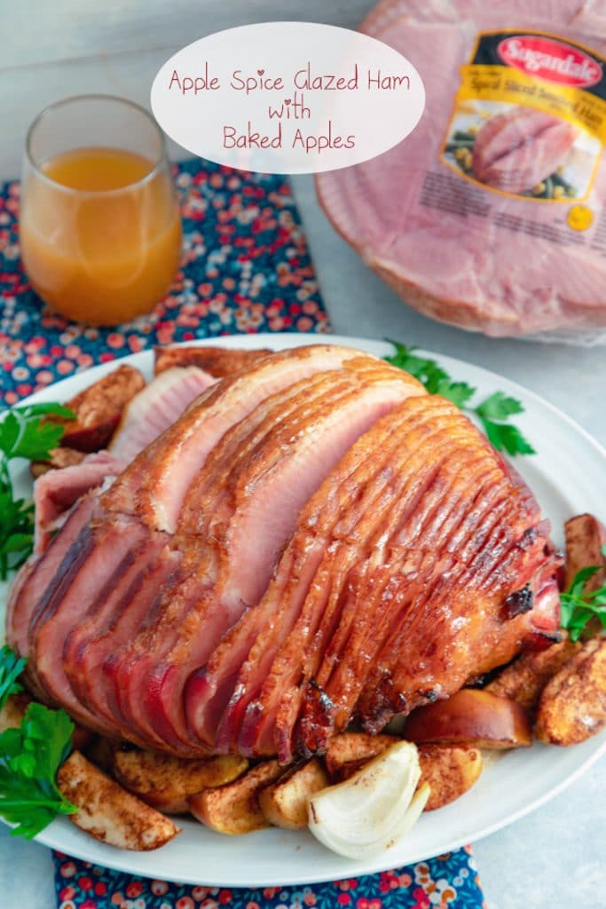 Overhead view of a sliced apple spice glazed ham in a spiral on a platter surrounded by baked apples with a glass of apple cider and another ham in the background and recipe title at top