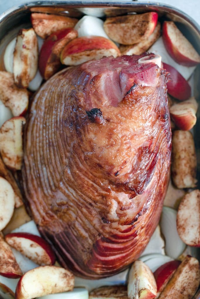 A vista de pájaro del jamón con especias de manzana cortado en espiral en su bandeja de hornear, rodeado de manzanas asadas y cebollas cortadas en rodajas