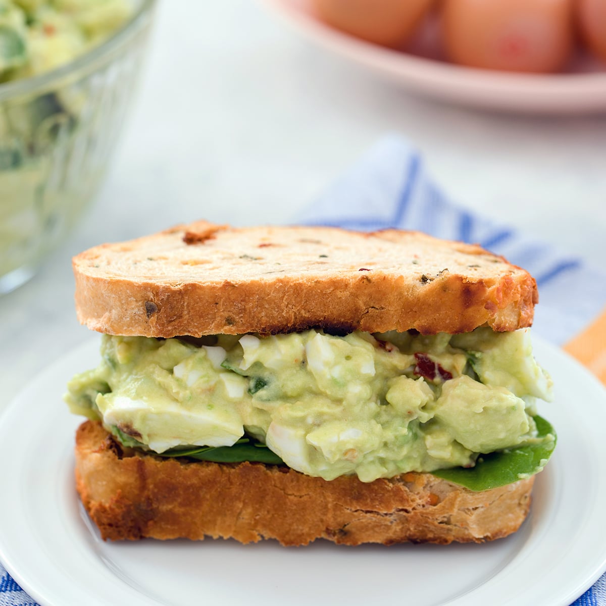 Head-on closeup view of a sandwich filled with avocado egg salad with spinach.