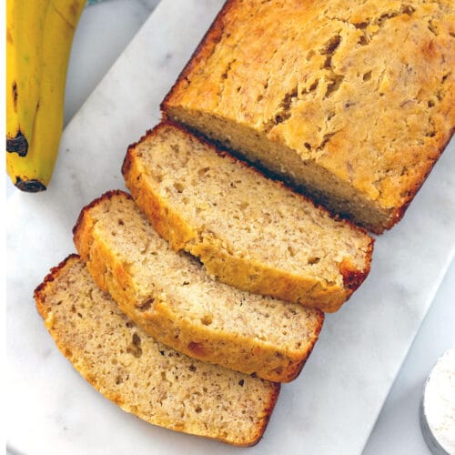 Slices of banana bread taken out of a loaf with bananas in background.