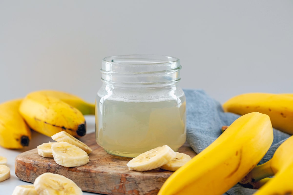 Head-on landscape view if a small jar of banana syrup on a wooden board with bananas all around.