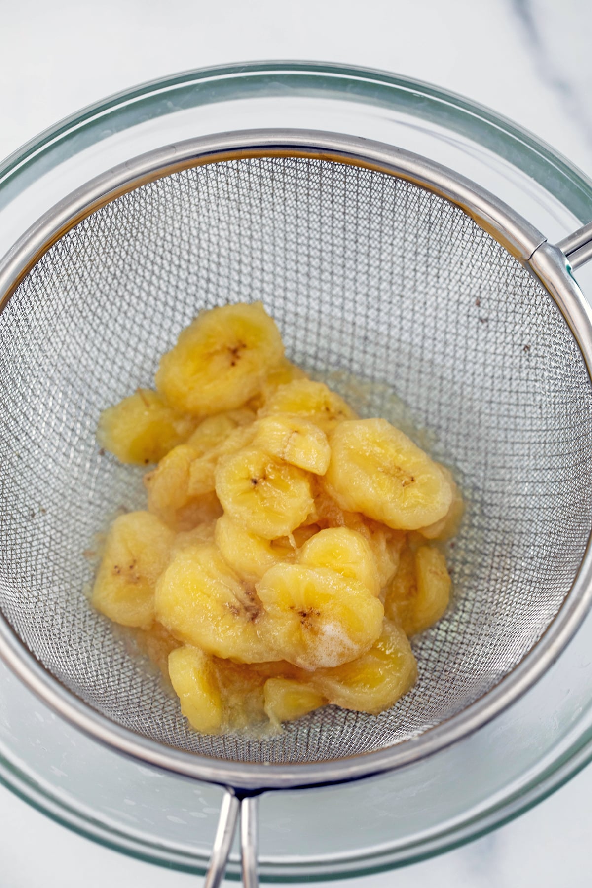 Mushed bananas being strained out of syrup in fine mesh sieve.