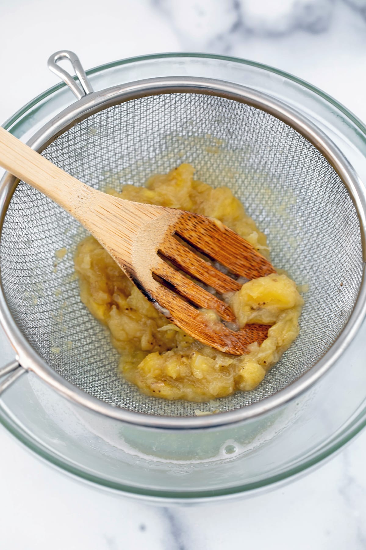 Wooden spoon mashing banana slices in fine mesh sieve.
