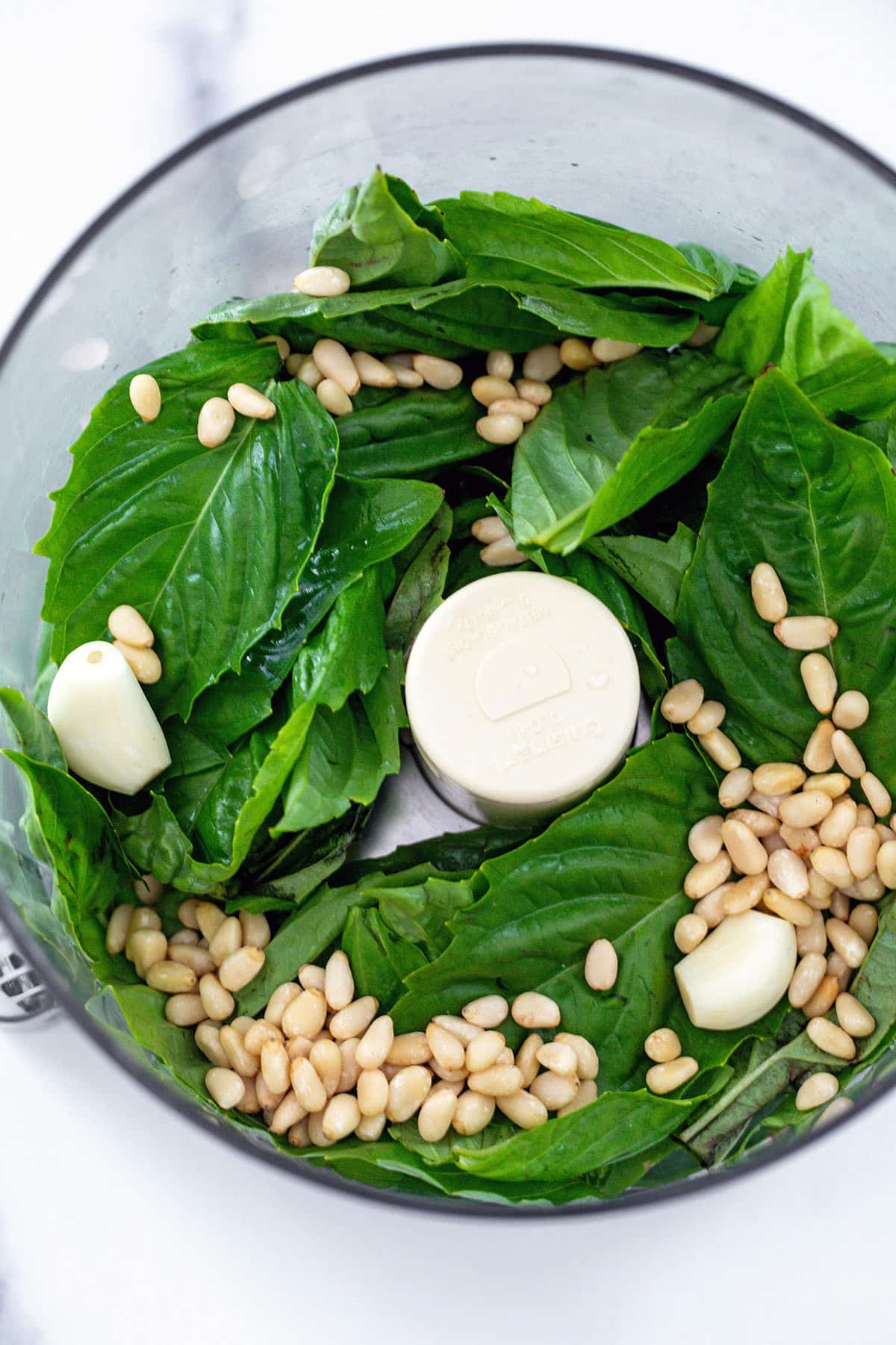 Fresh basil, pine nuts, and garlic cloves in food processor bowl.