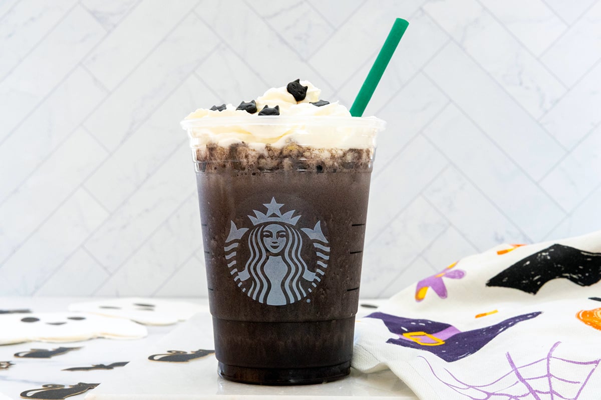 Landscape head-on view of Black Cat Frappuccino with whipped cream, cat sprinkles, and green straw in Starbucks cup.