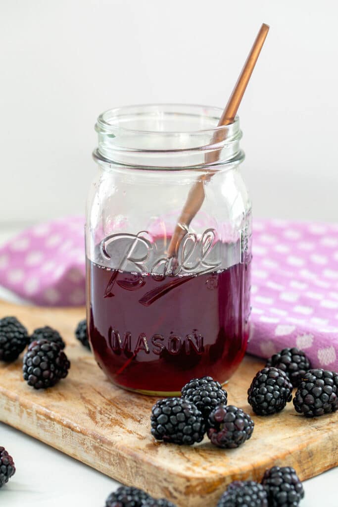 Large mason jar of blackberry simple syrup with spoon in it and blackberries all around.
