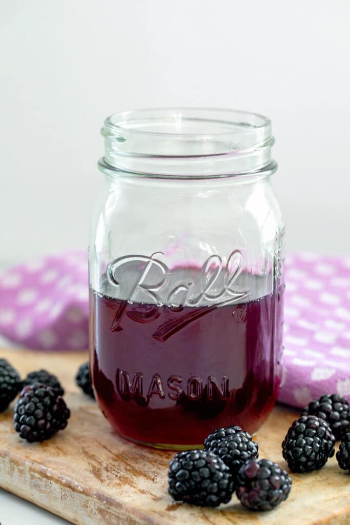 Head-on view of a mason jar of blackberry syrup on a wooden board with fresh blackberries.