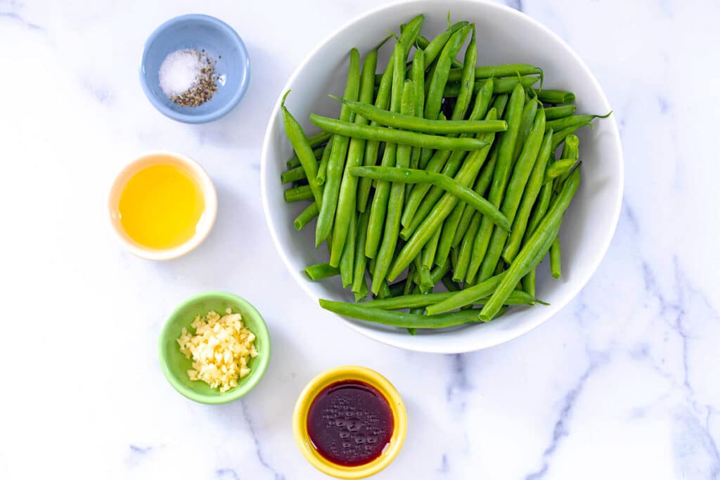 Fresh green beans in a bowl with small bowls of olive oil, salt and pepper, minced garlic, and soy sauce.