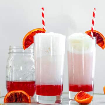 Head-on view of two blood orange ice cream sodas with jar of blood orange syrup in background.