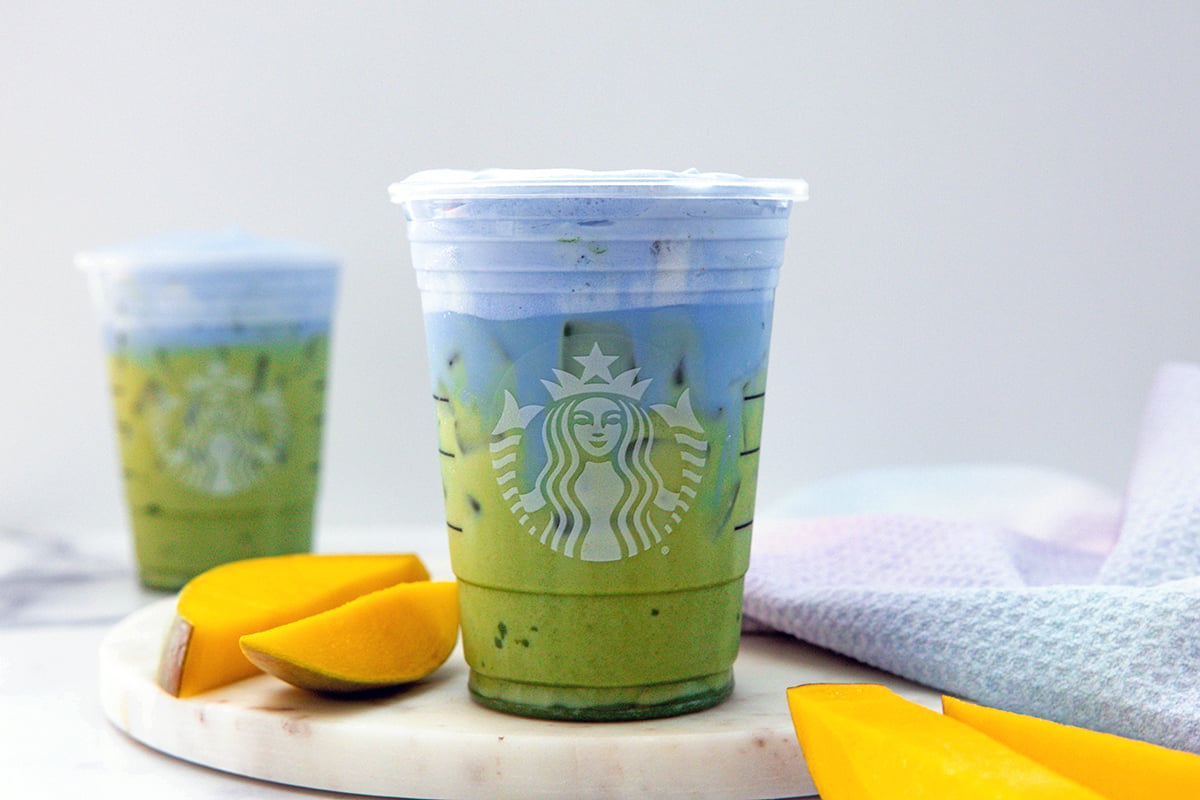 Landscape head-on view of a Blue Coconut Matcha with ice in a Starbucks cup with second drink in background and sliced mango all around.