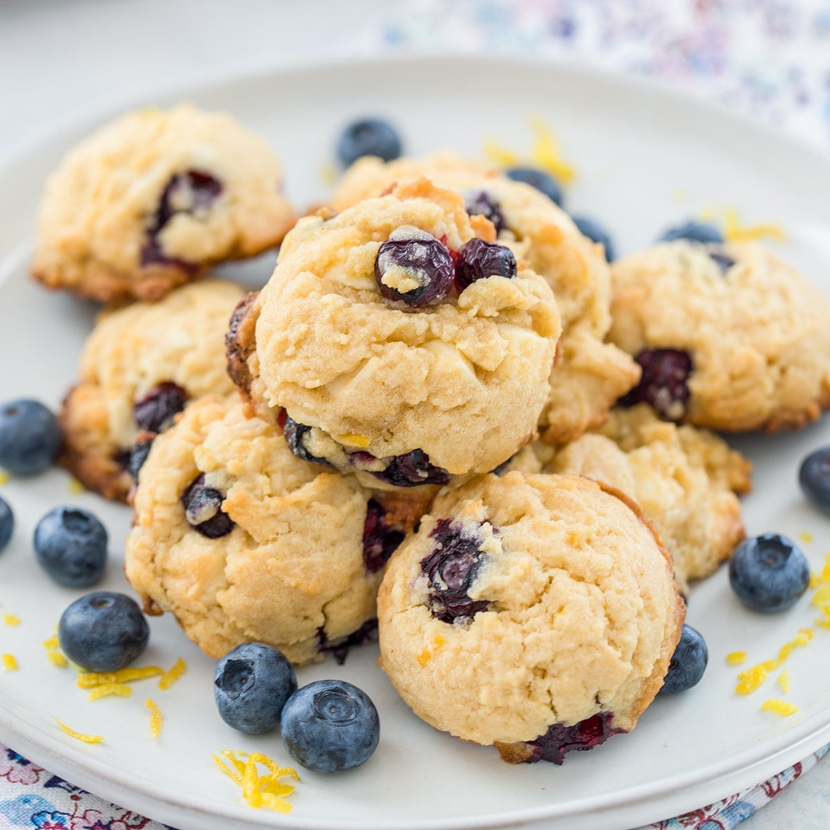 Blueberry, lemon, white chocolate chunk cookies on a plate with fresh blueberries and lemon zest all around.