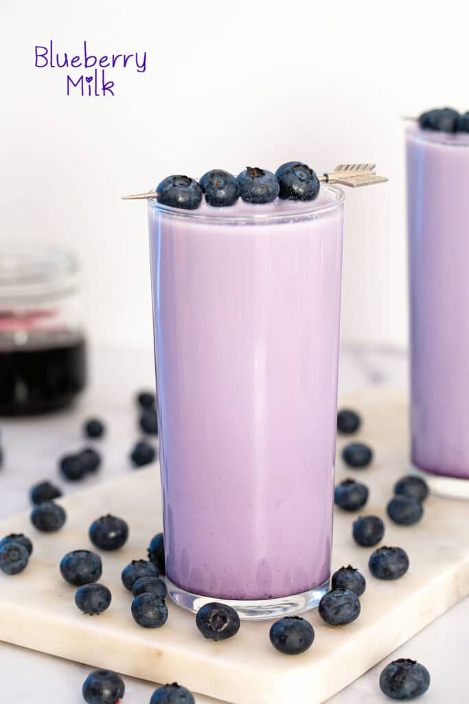 Head-on view of a glass of blueberry milk with blueberries all around and on garnish and recipe title at top.