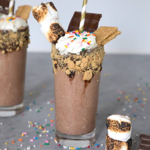 Closeup view of a bourbon s'mores milkshake with graham cracker rim and marshmallow, sprinkles, and chocolate bar toppings.
