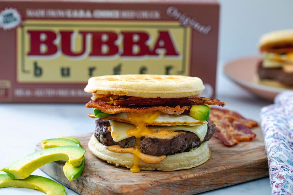 Waffle breakfast burger on a wooden board with avocado and bacon on the side and box of BUBBA burgers in the background.