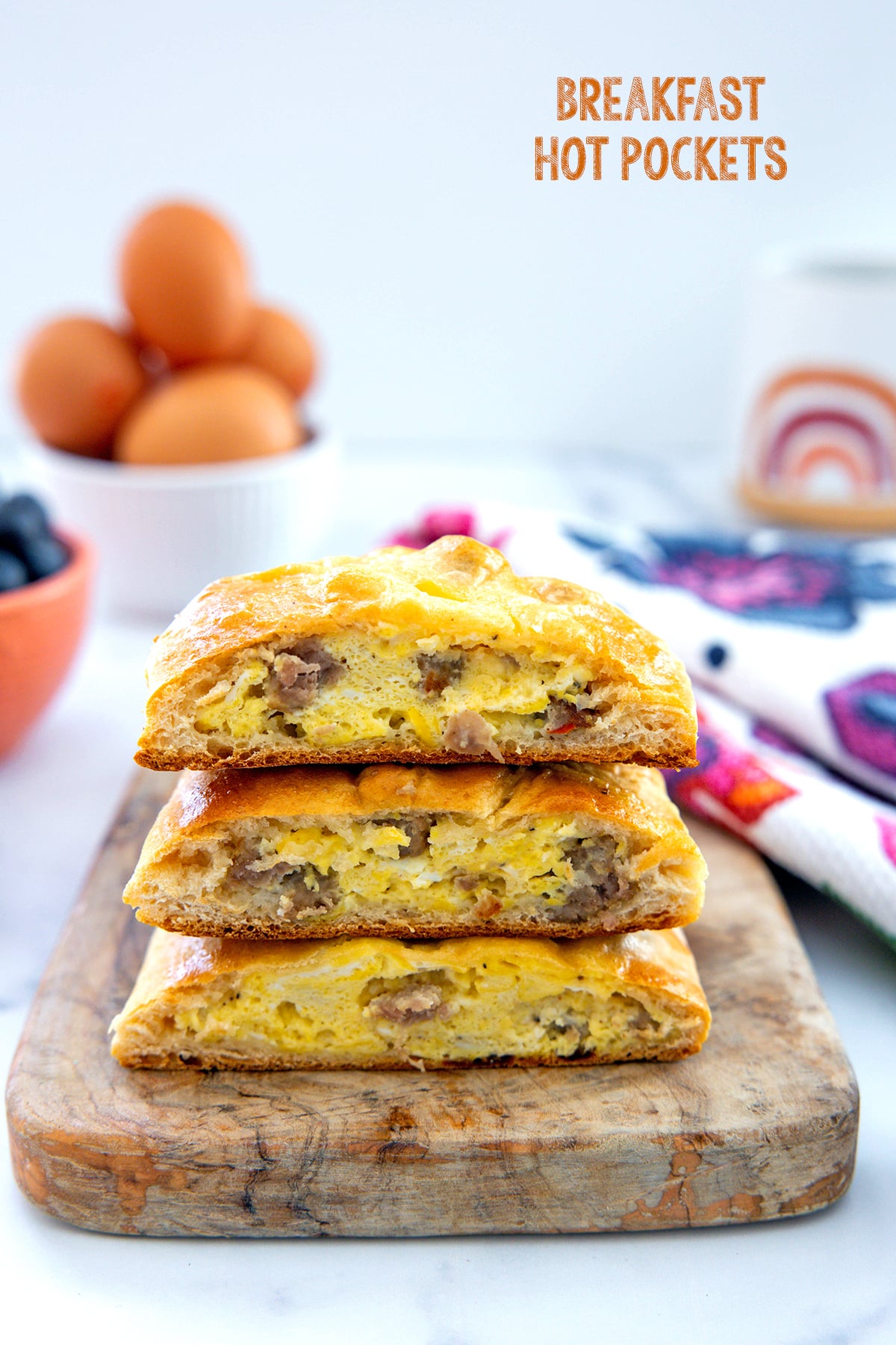 Head-on view of a stack of breakfast Hot Pockets filled with scrambled eggs and sausage with bowl of eggs and coffee cup in background and recipe title at top.
