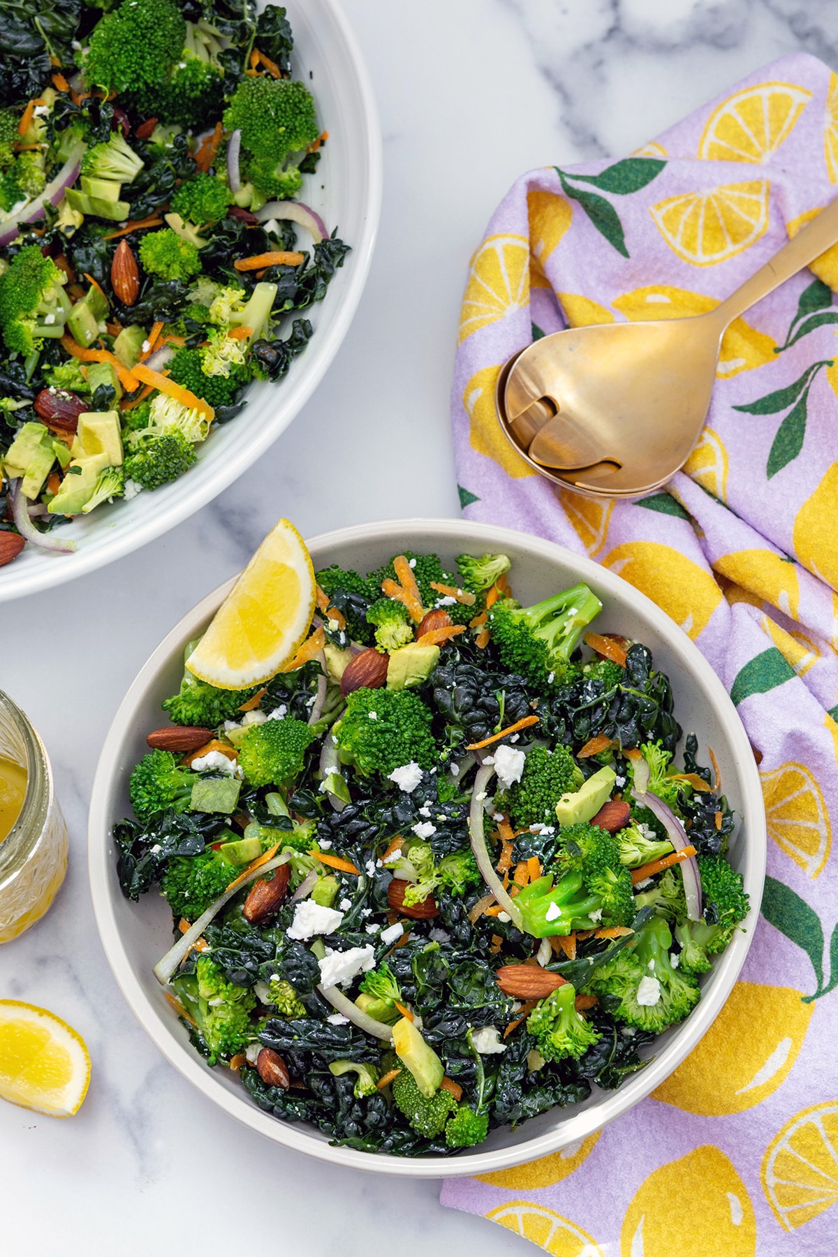 Bird's eye view of a bowl of broccoli kale salad with serving bowl and servings spoons in background.