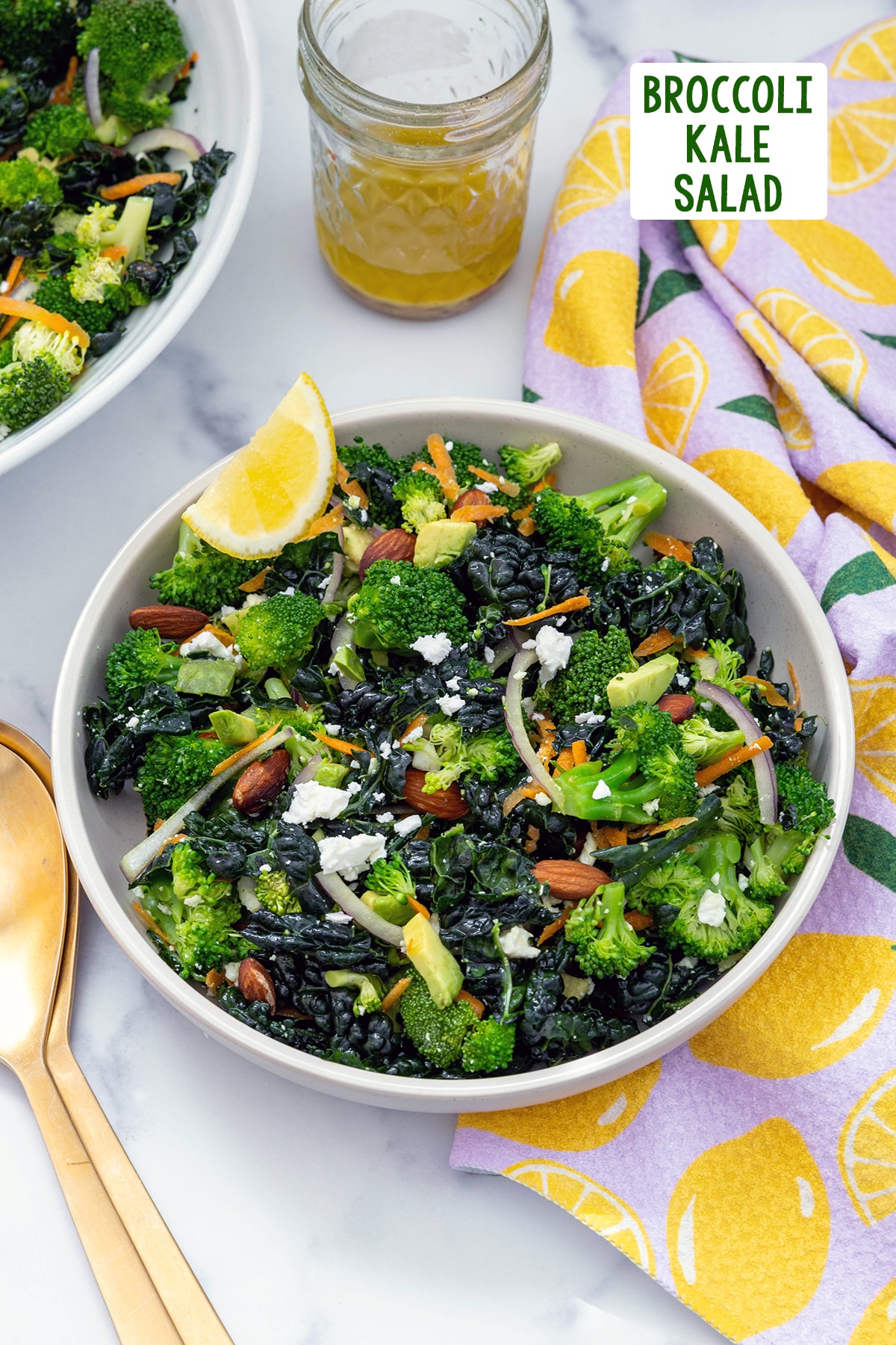 Broccoli kale salad with red onion, almonds, and a lemon wedge in a bowl with jar of dressing and recipe title at top.