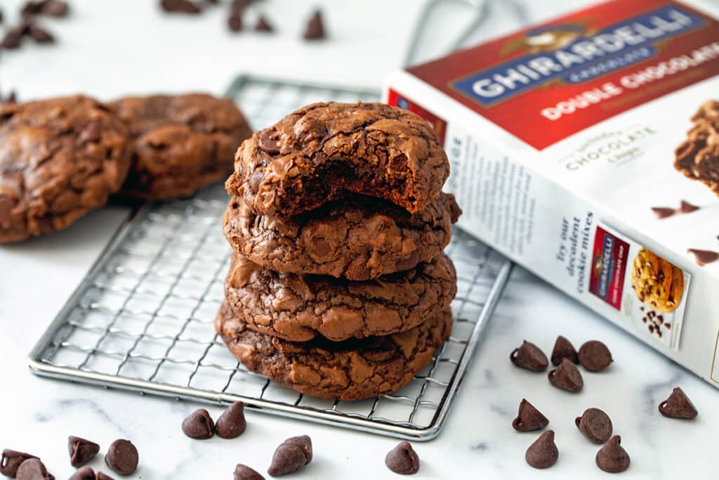 Landscape photo featuring a stack of brownie mix cookies, the top one having a bite out of it. There's cookies in the background, along with chocolate chips and a box of brownie mix.