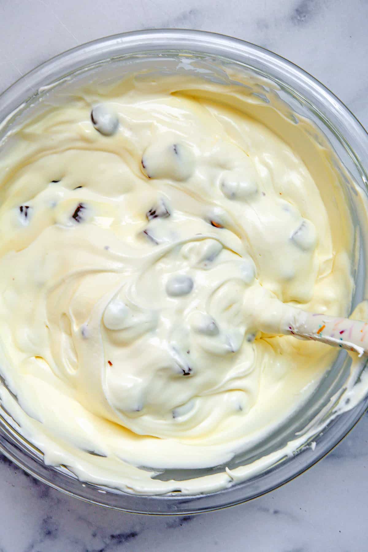Sweetened condensed milk, whipped cream, and chocolate candies mixed in with spatula in bowl.