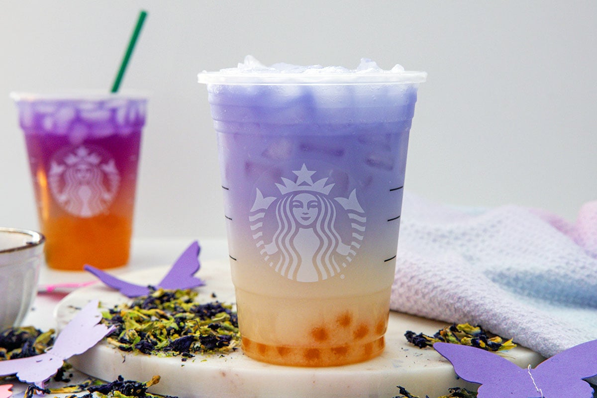 Landscape head-on view of a Butterfly Drink in a Starbucks cup with Tropical Butterfly Refresher in background and dried blue flowers all around.