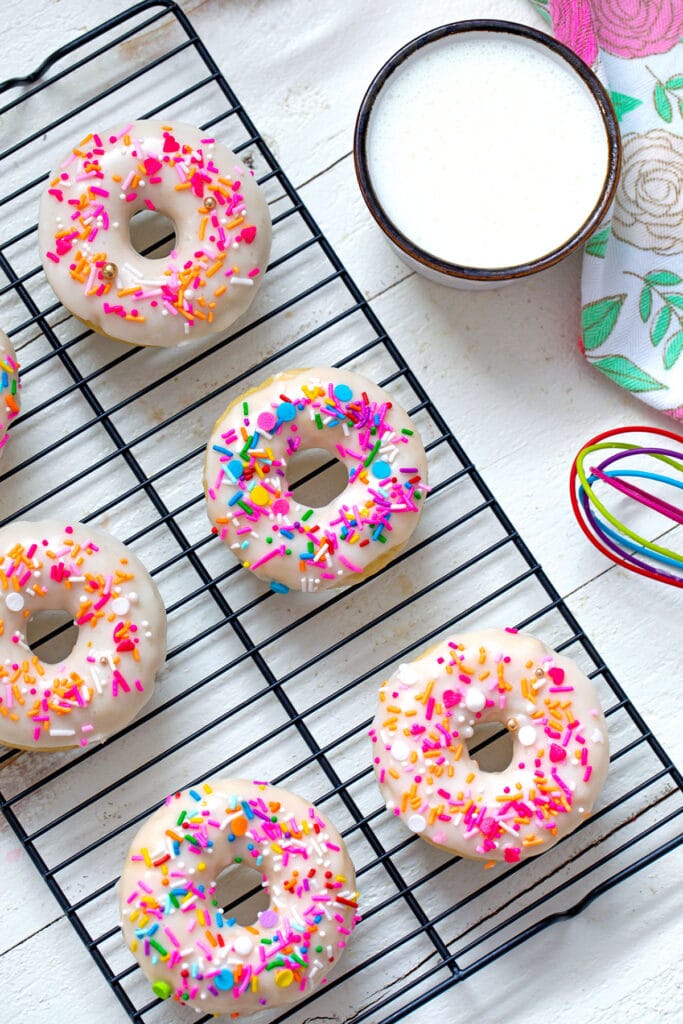 Overhead view of sprinkle topped buttermilk donuts on a baking rack with bowl of icing and whisk in background.