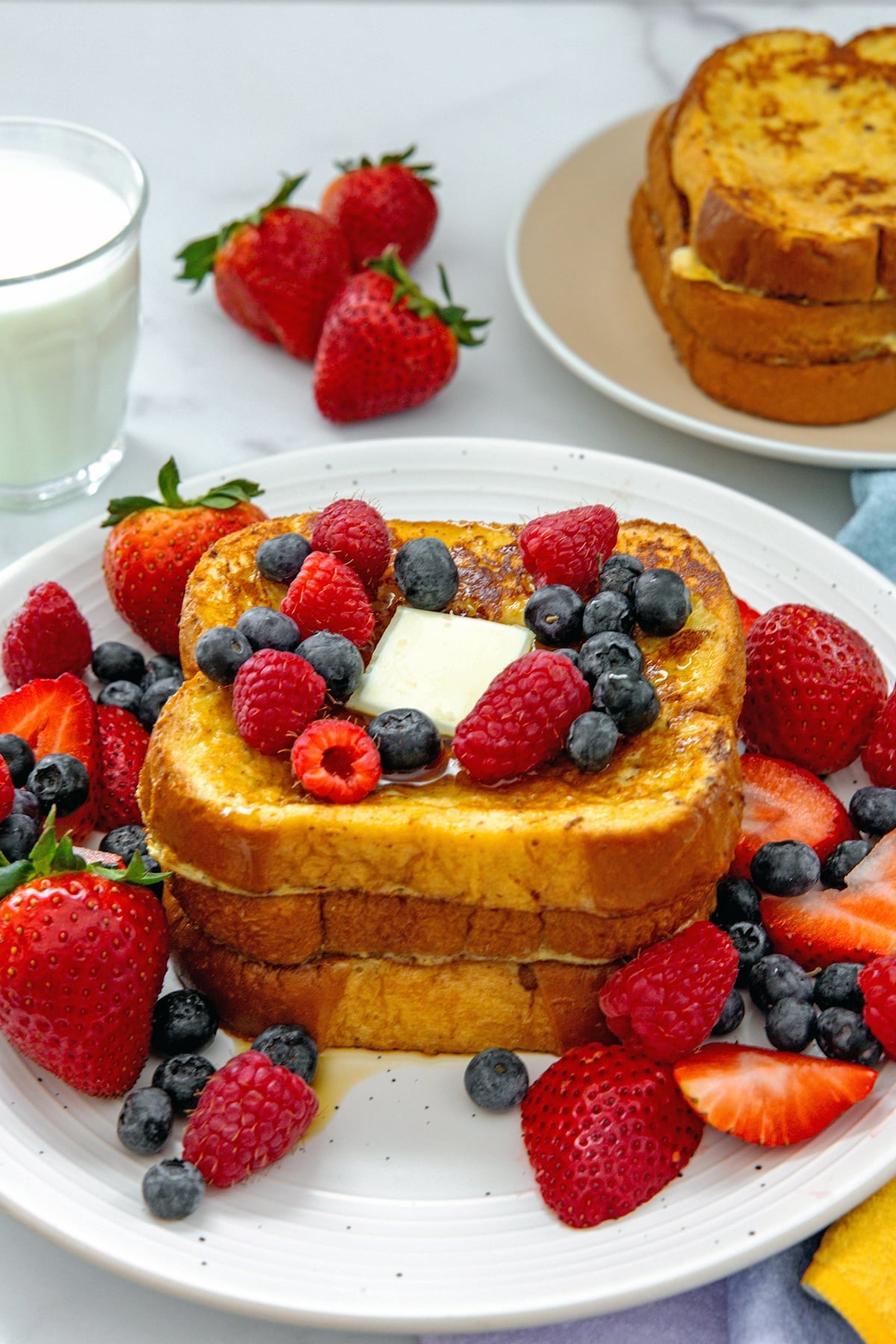 Stack of brioche buttermilk french toast topped with pat of butter and berries with second stack and glass of buttermilk in background.