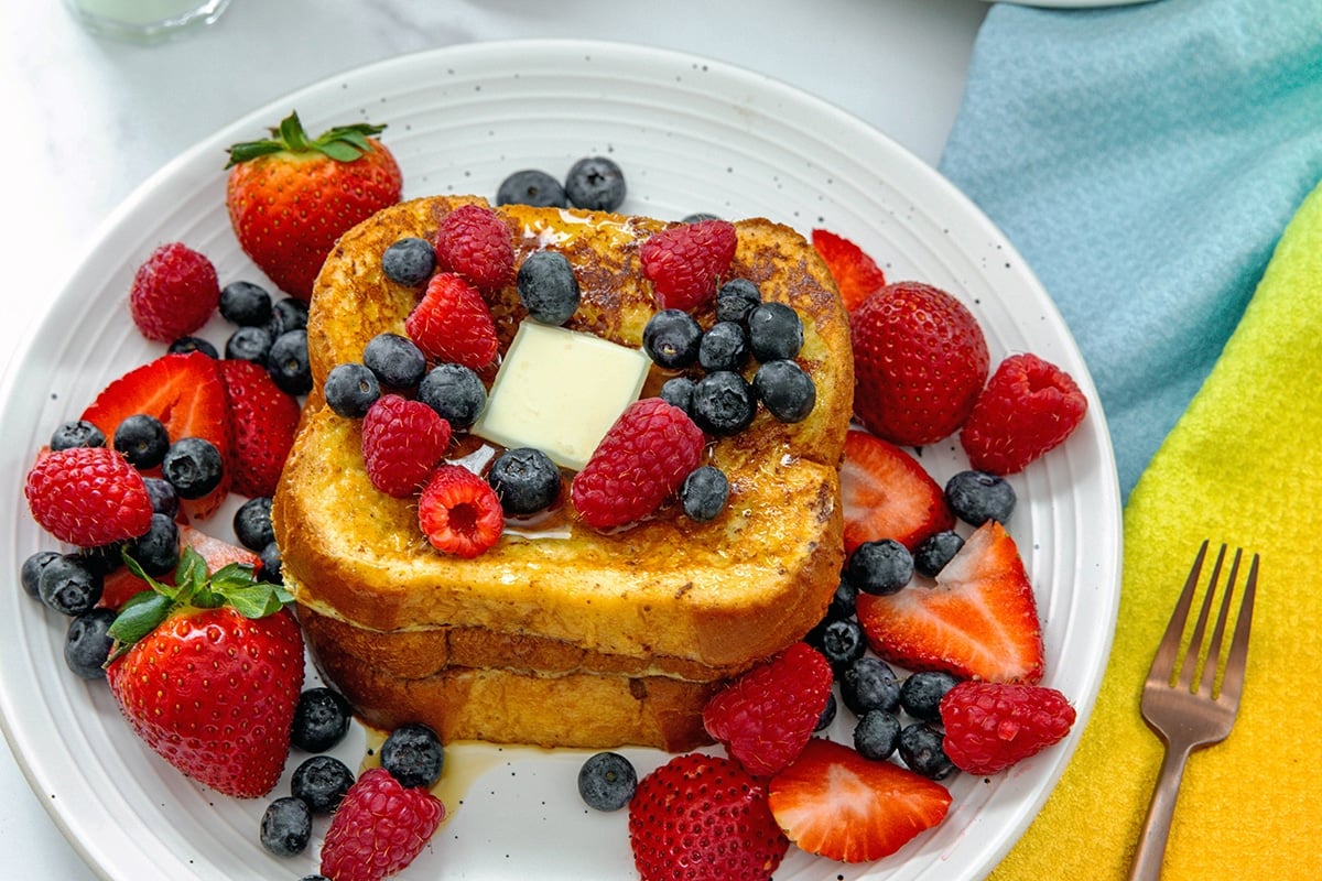 Overhead landscape view of a stack of brioche buttermilk french toast topped with pat of butter and berries.