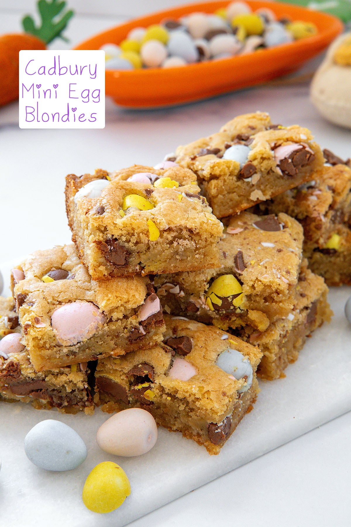 Cadbury Mini Egg blondies in a stack on a marble platter with candies all around and recipe title at top.