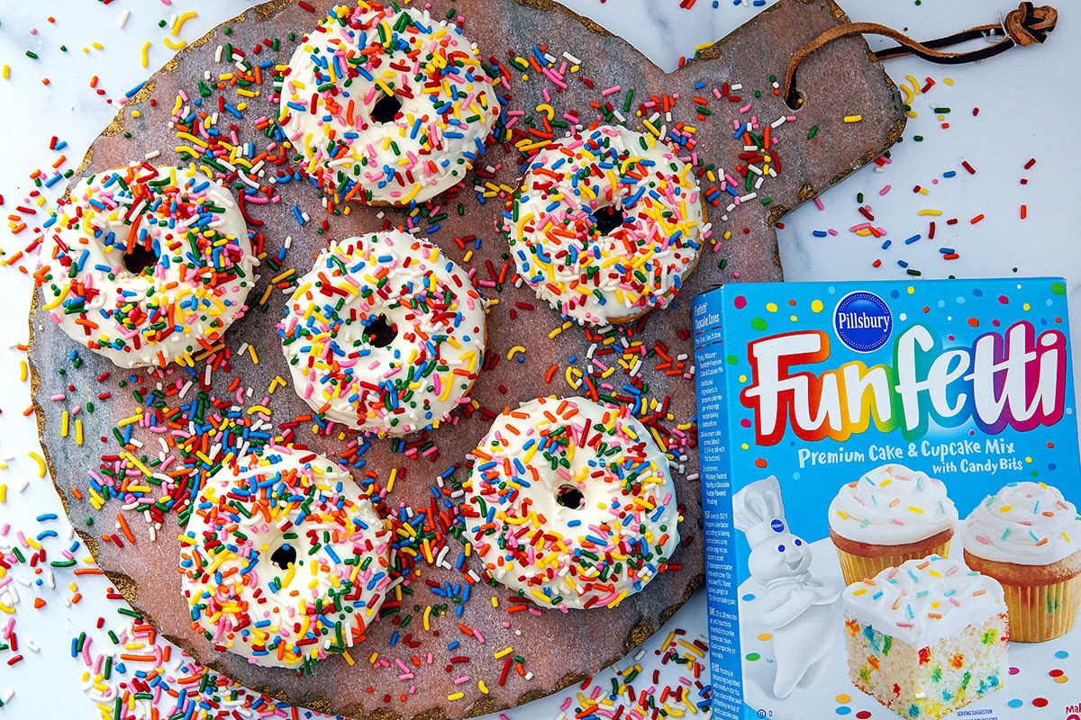 Landscape overhead view of cake mix donuts with rainbow sprinkles and box of Funfetti cake mix.