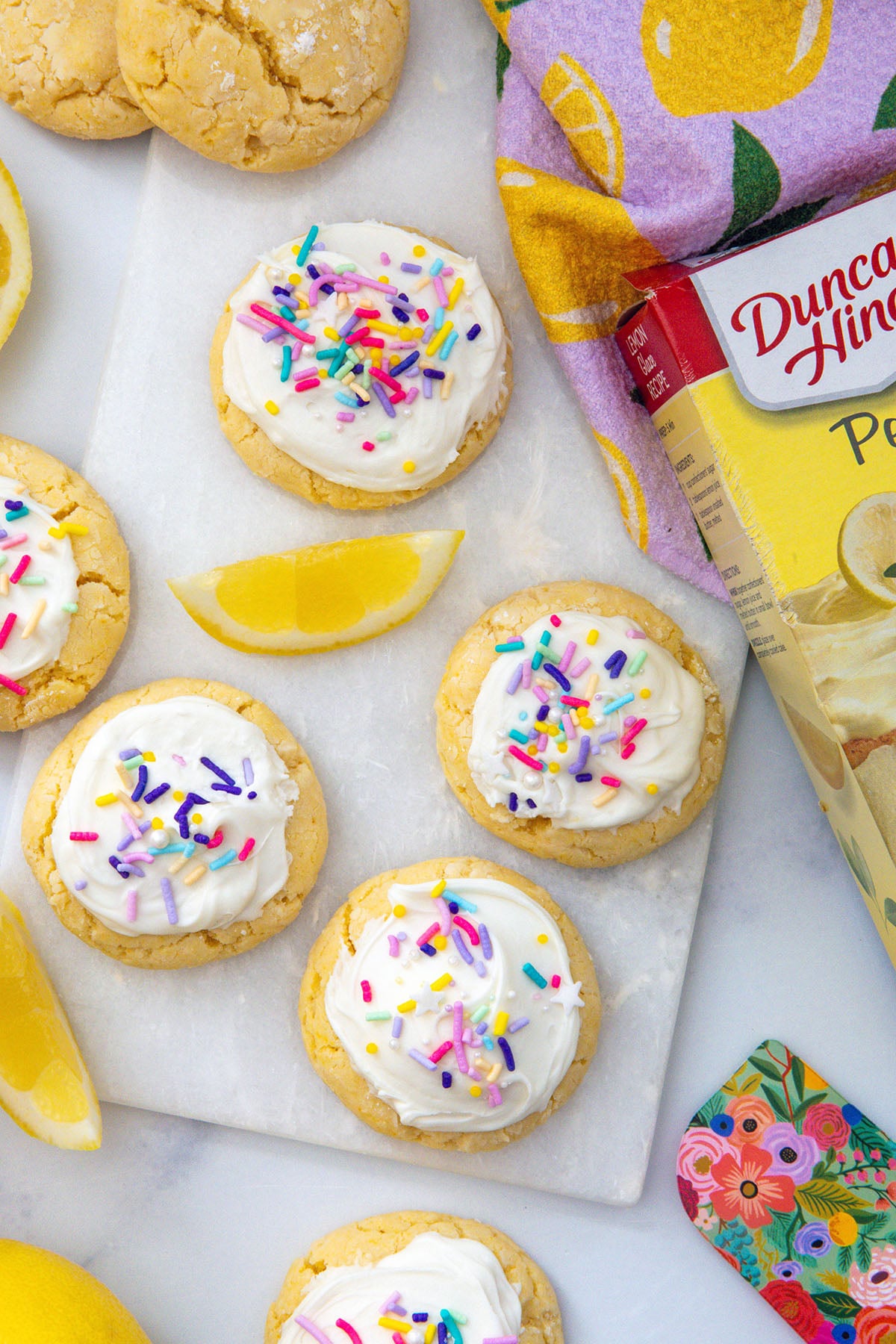 Bird's eye view of frosted lemon cake mix cookies with lemon wedges all around and box of cake mix on the side.
