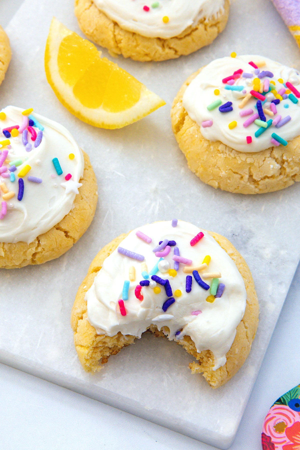 Closeup view of lemon cake mix cookies with frosting and sprinkles on a marble platter with one cookie with a bite taken out of it.