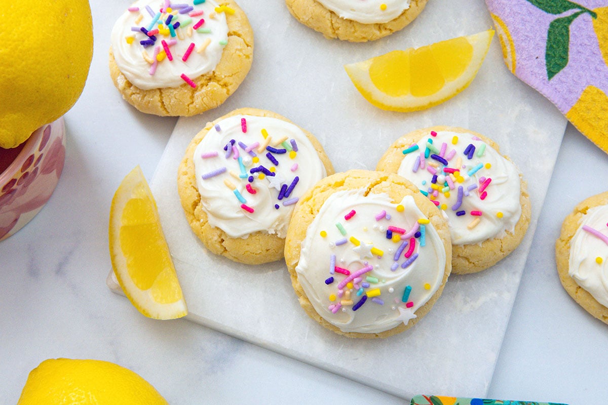Landscape view of lemon cake mix cookies with frosting and sprinkles with lemon wedges all around.