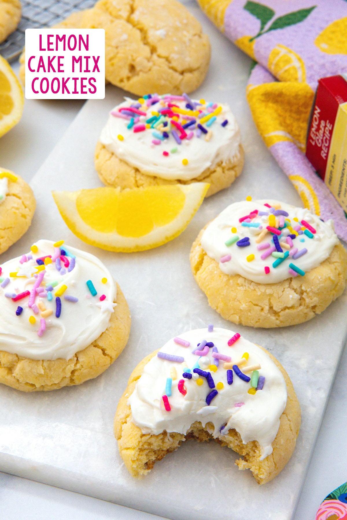 Overhead view of frosted lemon cake mix cookies on a marble tray with a bite out of one, lemon wedges all around, and recipe title at top.