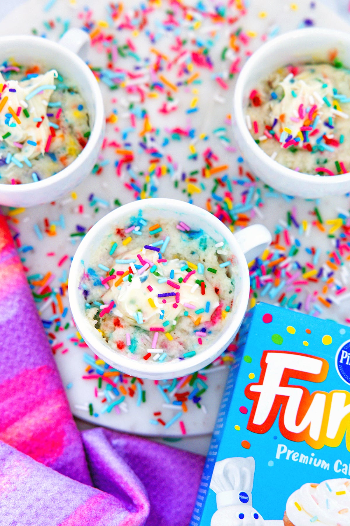 Overhead view of multiple mug cakes with sprinkles all around and box of funfetti cake mix.