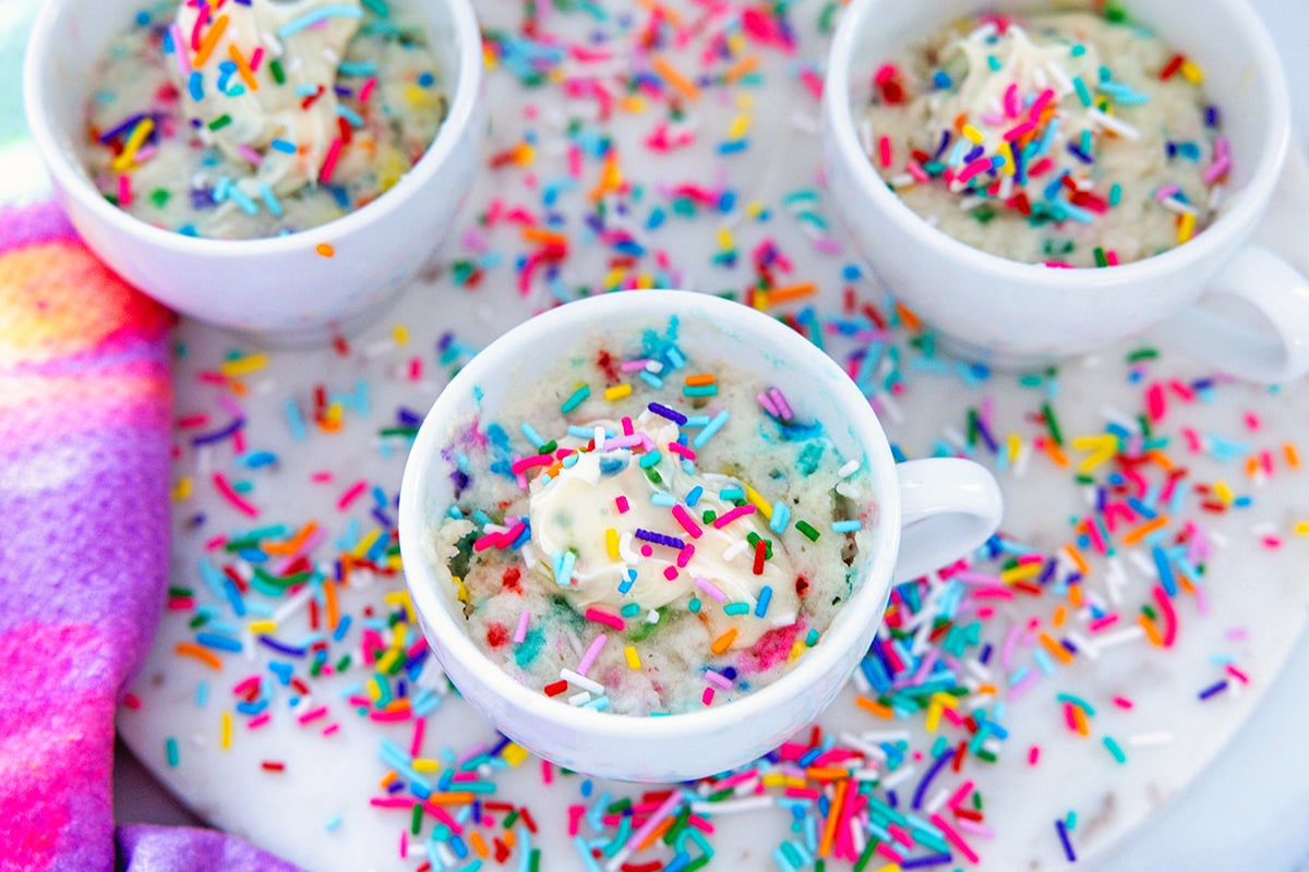 Landscape overhead view of multiple white mugs with cake mix mug cake and dollops of frosting with sprinkles all around.